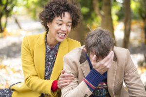 Two people sit on a park bench. One covers their face and hunches over with sadness; the other touches their arm and smiles, trying to cheer them up.