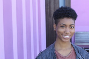 A person with short hair is smiling into the camera with a striped purple backdrop behind them.