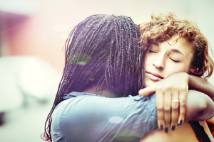 Two friends are hugging one another – one with their face turned away from the camera, the other with their eyes closed and facing the camera.