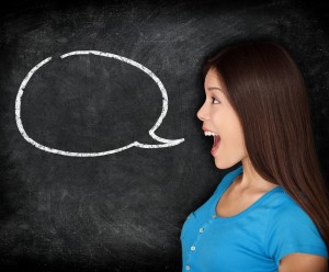 Person in a blue shirt with long, dark hair standing in front of a chalk board that has a speech bubble drawn on it